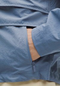 Vaaleansininen takki, jossa on tekstuuripinta, varustettuna vetoketjullisella taskulla ja tiiviillä hihansuilla. Materiaali näyttää olevan kevyt ja ryppyilemätön.