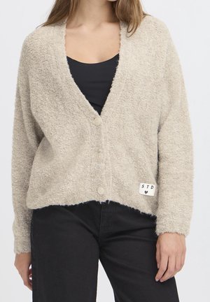 Femme portant un cardigan beige duveteux boutonné par-dessus un haut noir et un pantalon noir, debout devant un fond clair uni.
