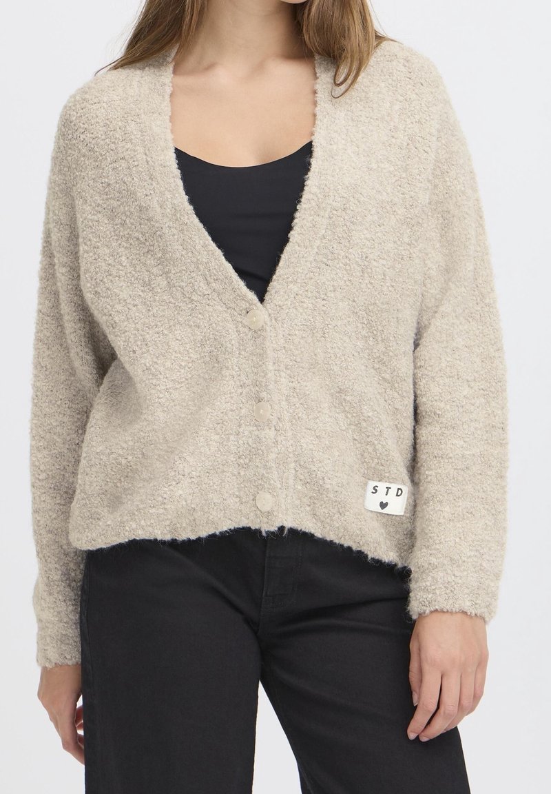 Femme portant un cardigan beige duveteux boutonné par-dessus un haut noir et un pantalon noir, debout devant un fond clair uni.