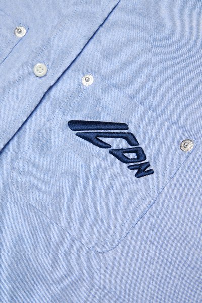 Camicia azzurra con un tessuto textured, caratterizzata da bottoni bianchi e un logo ricamato blu navy sulla tasca.