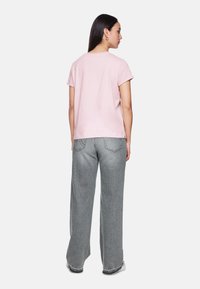 Frau, die von hinten schaut, trägt ein hellrosa T-Shirt mit kurzen Ärmeln, graue weit geschnittene Jeans und weiße Sneakers vor einem weißen Hintergrund.