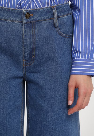 Ljusblå denimjeans med synlig sömnad och en framficka. En hand vilar på sidan och visar en slät yta.