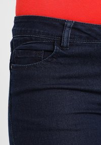 Detailní záběr na tmavě modré džíny v pase a kapsách, kombinované s zakasaným jasně červeným topem.