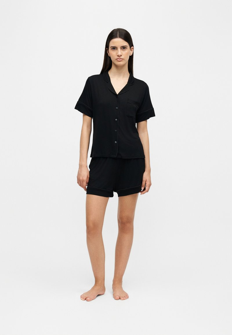 Femme debout pieds nus portant un haut de pyjama noir à manches courtes boutonné et un short assorti sur un fond clair uni.