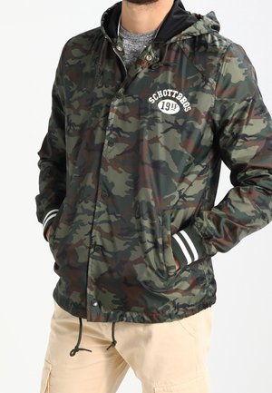 Homme portant une veste camouflage verte et marron avec un logo blanc sur la poitrine, des poignets rayés et un pantalon cargo beige, les mains dans les poches.