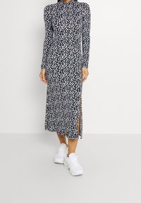 Långärmad midiklänning i svart med vita prickar, utrustad med hög urringning och sidoklipp, i sällskap med vita träningssneakers.