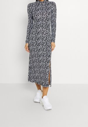Langszijde midi jurk in zwart met witte stippen, voorzien van een hoge hals en zijsplitten, gedragen met witte sportschoenen.