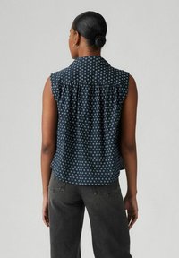 Blusa senza maniche di colore blu navy con motivo floreale bianco, dotata di colletto e dettaglio sul retro arricciato. Abbinata a jeans grigi a vita alta.