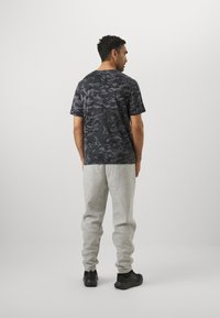 Sort camouflage-mønstret t-shirt med korte ærmer parret med lysegrå sweatpants. Modellen står med ryggen mod beskueren og har sorte sko på.