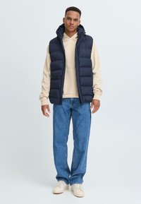 Marineblauer Steppweste mit hohem Kragen und vorderem Reißverschluss, getragen über einem hellbeigen Hoodie, kombiniert mit hellblauen Jeans und cremefarbenen Sneakern.