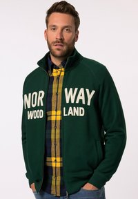 Green zip-up jacket with "NORWAY WOOD LAND" printed in white. Worn over a blue and yellow plaid shirt with a button-down collar.
