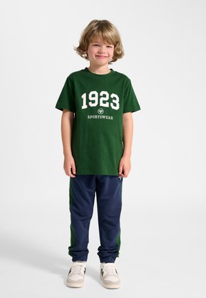 Smiling child with blonde hair wearing a green "1923 Sportswear" t-shirt, navy and green pants, and white sneakers standing against a plain background.