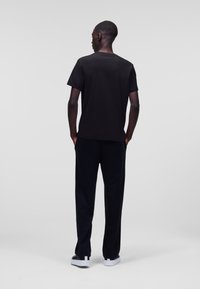 Zwarte katoenen T-shirt met ronde hals, gecombineerd met zwarte broek, gezien vanaf de achterkant. Glad textuur en rechte pasvorm. Witte schoenen.
