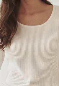 Ribbad krämfärgad topp med rundad halsringning. Utmärks av elastisk tyg och en nära passform, framhävt av subtil textur och linjer.