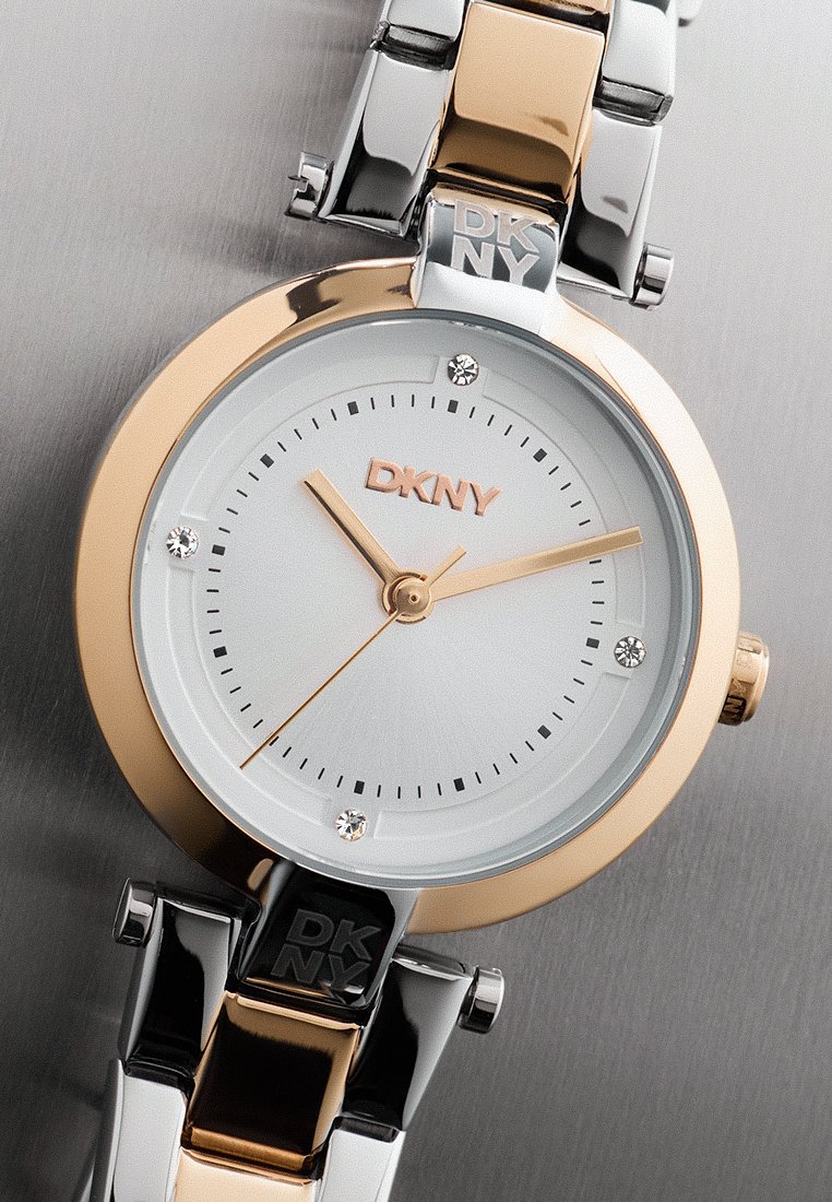 DKNY horloge met een ronde roségouden behuizing, witte wijzerplaat met kristallen uurmarkeringen, gouden wijzers en een twee-tone metalen armband.