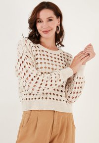 Suéter de punto en beige claro con patrón de agujeros abiertos, escote redondo y puños acanalados. Combinado con pantalones color beige.