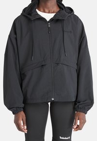 Giacca nera leggera con cerniera frontale, cappuccio regolabile e polsini elasticizzati; presenta un design semplice e lineare senza motivi visibili.