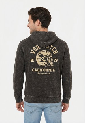 Hombre con sudadera negra desgastada con capucha, con gráfico de calavera y casco y texto "Von Dutch California Motorcycle Club" en la parte trasera, pantalones vaqueros azul claro, de espaldas.
