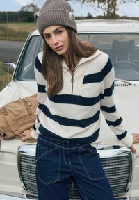 Jeune femme en pull rayé et bonnet tricoté appuyée contre une voiture vintage blanche avec un sac à dos beige posé sur le capot, dans un cadre rural en plein air.