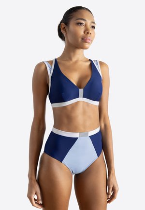 Frau trägt einen blau und hellblau hoch taillierten Bikini mit weißem Besatz und schaut selbstbewusst seitlich vor einfarbigem Hintergrund.