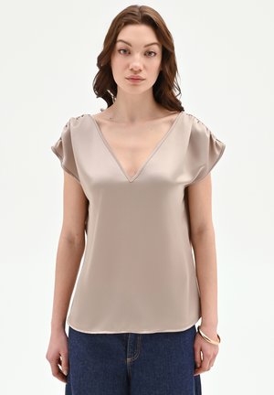 Donna che indossa una camicia beige in raso a maniche corte con scollo a V e pantaloni di denim blu, in piedi contro uno sfondo chiaro uniforme.