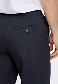 Pantaloni blu realizzati in tessuto liscio, con una tasca posteriore dotata di un bottone blu e una vita standard.