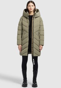 Abrigo acolchado verde oliva con capucha, con costuras en forma de chevrón, dos bolsillos delanteros y cierre de cremallera. Llevado sobre leggings negros y botas.