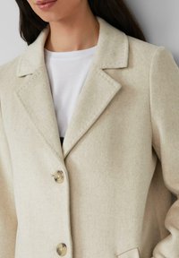 Manteau en mélange de laine crème avec col cranté, fermeture à un bouton et détails de couture visibles. Accompagné d'une chemise blanche en dessous.