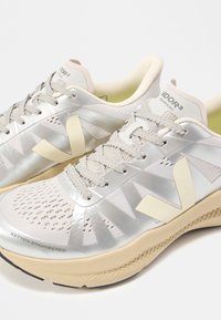 Ανοιχτό μπεζ και εκρού δαντελωτά παπούτσια τρεξίματος με χοντρές μαλακές σόλες και μεγάλα λογότυπα "V" στις πλευρές, τοποθετημένα σε λευκή επιφάνεια.