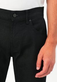 Close-up of a person wearing black denim jeans with a button and pocket visible, alongside their right hand resting by their side.