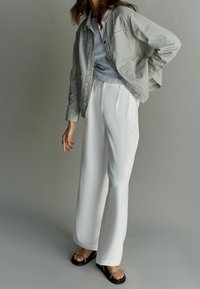 Gestreepte lichtgrijze jas over een blauw topje, gecombineerd met witte wijde broekspijpen en zwarte sandalen met een dunne riem.