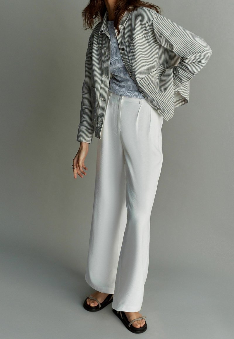 Gestreepte lichtgrijze jas over een blauw topje, gecombineerd met witte wijde broekspijpen en zwarte sandalen met een dunne riem.
