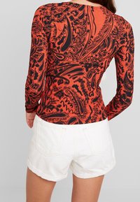 Langarmshirt in leuchtendem Orange mit schwarzen Paisley-Mustern. Kombiniert mit weißen Shorts, die ein figurbetontes und strukturiertes Design aufweisen.