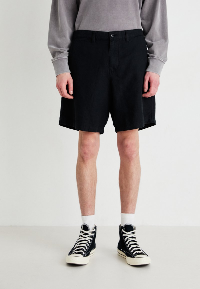 Schwarze Baumwollshorts mit geradem Schnitt, ausgestattet mit einem Knopfverschluss und seitlichen Taschen, kombiniert mit hohen schwarzen Sneakern.