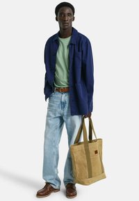 Marineblause über einem hellgrünen T-Shirt, hellblaue Jeans, braune Schuhe und eine braune sowie beige Tragetasche mit grünen Trägern.