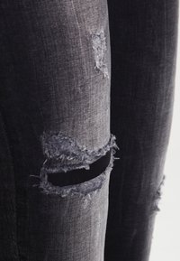 Jean en denim gris foncé présentant des zones usées avec des bords effilochés, mises en valeur par des déchirures prononcées et des textures de tissu contrastées.