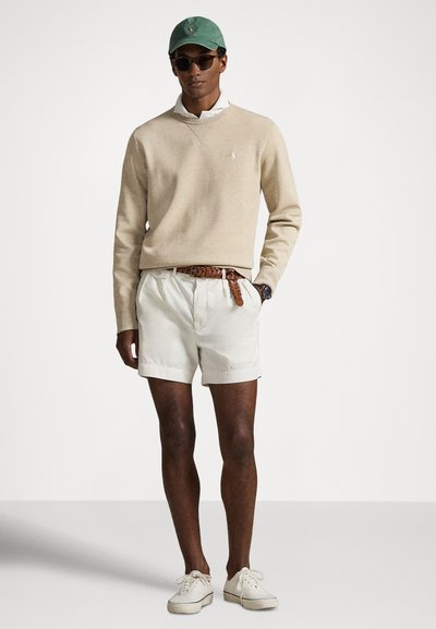 Μπεζ cotton πουλόβερ με ελαφρύ V-λαιμόκοψη, κρεμ σορτς, καφέ πλεκτή ζώνη, πράσινο καπέλο και σκούρα γυαλιά ηλίου· casual ντύσιμο με ελαφριά αθλητικά παπούτσια.