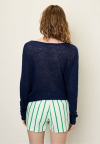 Tamsiai mėlynas lengvas megztinis su plačiu iškirpimu ir ilgomis rankovėmis, derintas su baltais šortais su žaliais vertikaliais dryžiais.