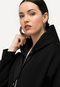 Hoodie noir avec fermeture éclair et capuche à cordon de serrage ; présente une texture de tissu lisse et du matériel en métal. Porté avec un haut blanc en dessous.