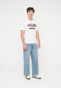 Λευκό μπλουζάκι με τυπωμένο λογότυπο και κείμενο, σε συνδυασμό με ανοιχτό μπλε τζιν παντελόνι με φαρδιά μπατζάκια και γκρι αθλητικά παπούτσια με μαύρες ρίγες. Απλός, καθημερινός σχεδιασμός.