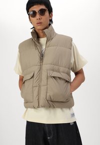 Beige padded vest with high collar, front zip, two buttoned pockets. Worn over a light cream shirt with short sleeves and dark jeans.