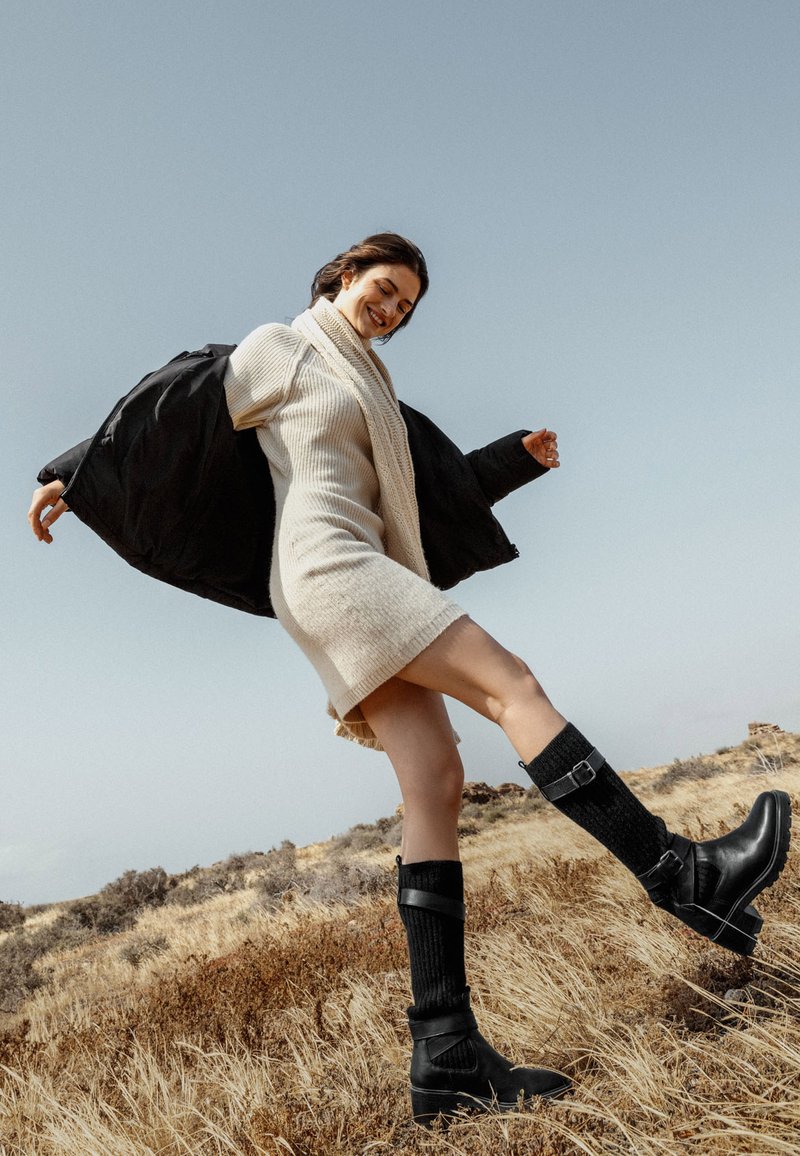 Chaqueta negra acolchada, vestido crema de canalé, botas negras de punto y cuero hasta la rodilla con detalles de hebilla, de pie sobre un fondo de terreno natural.