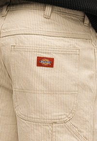 Ποτζέτο μπεζ και λευκής ριγέ παντελονιού με ένα κόκκινο έμβλημα λογότυπου Dickies ραμμένο πάνω από το ποτζέτο στην πίσω πλευρά του παντελονιού.