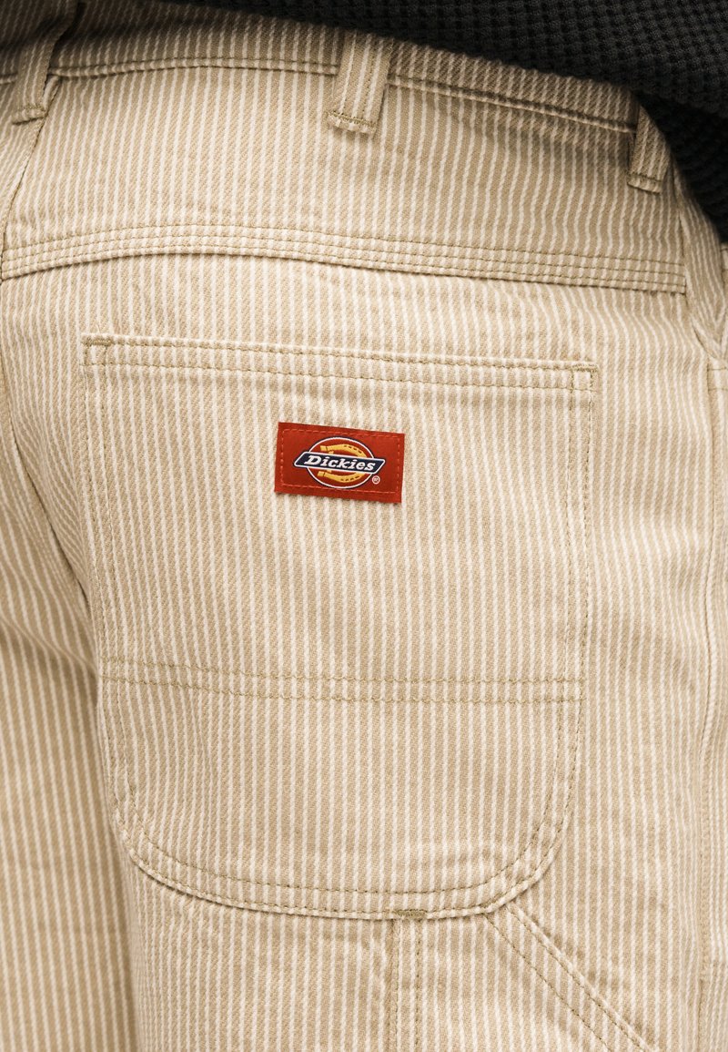 Beige en wit gestreepte broekzak met een rood Dickies-logo patch genaaid boven de zak aan de achterkant van de broek.