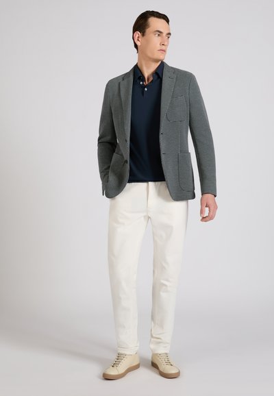 Hombre con blazer gris de estampado, camisa azul oscuro, pantalones blancos y zapatillas deportivas beige, de pie y mirando hacia la derecha sobre un fondo liso.