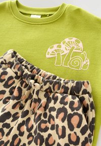 Zielona bluza z haftem w kształcie różowej muchomora i ślimaka, zestawiona z beżowymi dresami w czarno-rude wzory z leopardem i elastycznym pasem ze sznurkiem.