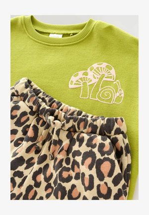 Zaļš krekls ar rozā sēņu un gliemeža izšuvumu, apvienots ar bēšām sporta biksēm, kas rotātas ar melnu un rūsu leoparda rakstu un ir ar auklu viduklī.