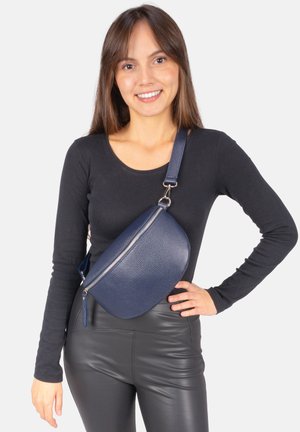 Marineblaue Leder-Umhängetasche mit abgerundeter Form, strukturierter Oberfläche, silbernem Reißverschluss und verstellbarem Riemen, getragen über einem schwarzen Oberteil.