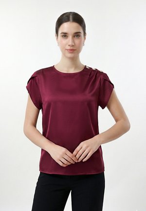 Jeune femme aux cheveux foncés portant un chemisier en satin bordeaux à manches courtes avec trois boutons dorés sur l'épaule gauche, debout devant un fond blanc.