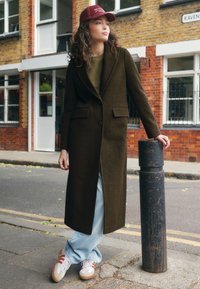 Long manteau vert olive avec un revers cranté, associé à un jean bleu ample et des baskets. Le mannequin porte une casquette bordeaux et pose contre un poteau de rue.
